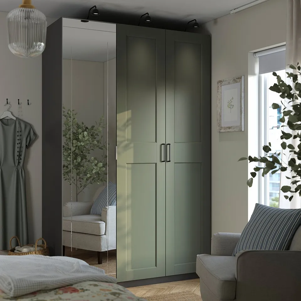 Modern bedroom a large, green PAX wardrobe with mirrored doors, standing tall beside a cosy sofa and next to a window, with subtle grey-green walls.