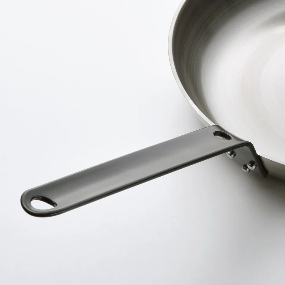 A close-up of a carbon steel pans handle and part of its base. The handle is dark grey with riveted joints and the pan has a polished surface.