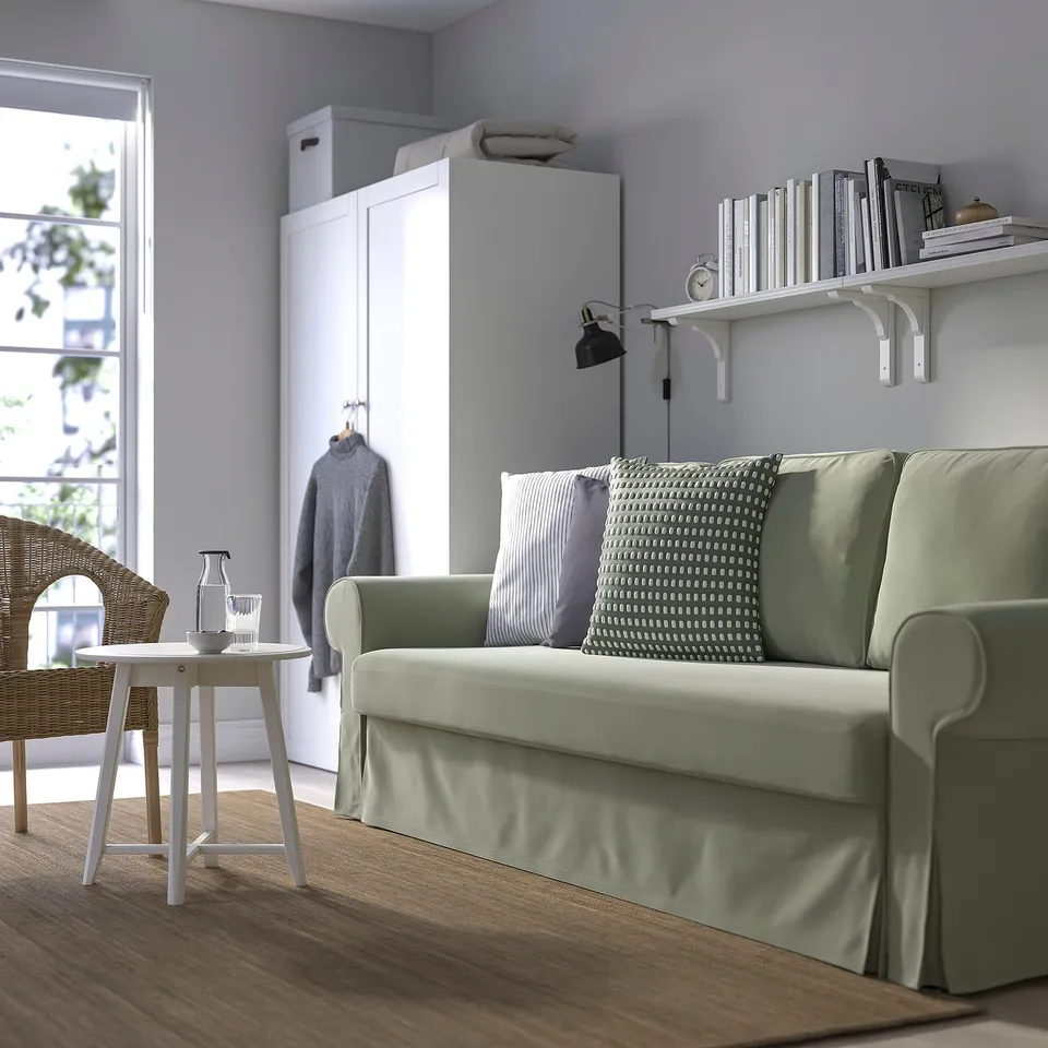 Green VRETSTORP sofa-bed in living room, next to wardrobe, under book shelf. Minimalist design with wood floors, white table, and chair.
