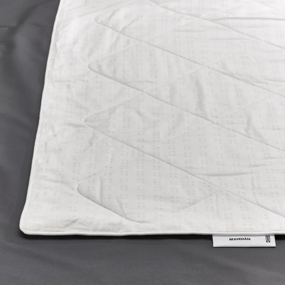 Clean, white duvet with elegant stitching, neatly laid on a grey surface. Textured cotton cover with a brand tag labelled IKEA and MJUKDÅN.