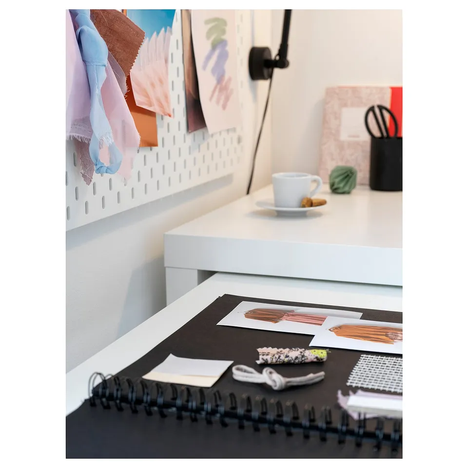 Open notebook with fabric swatches, sketches, coffee cup, and cleaning supplies on desk. Pegboard with textiles above.