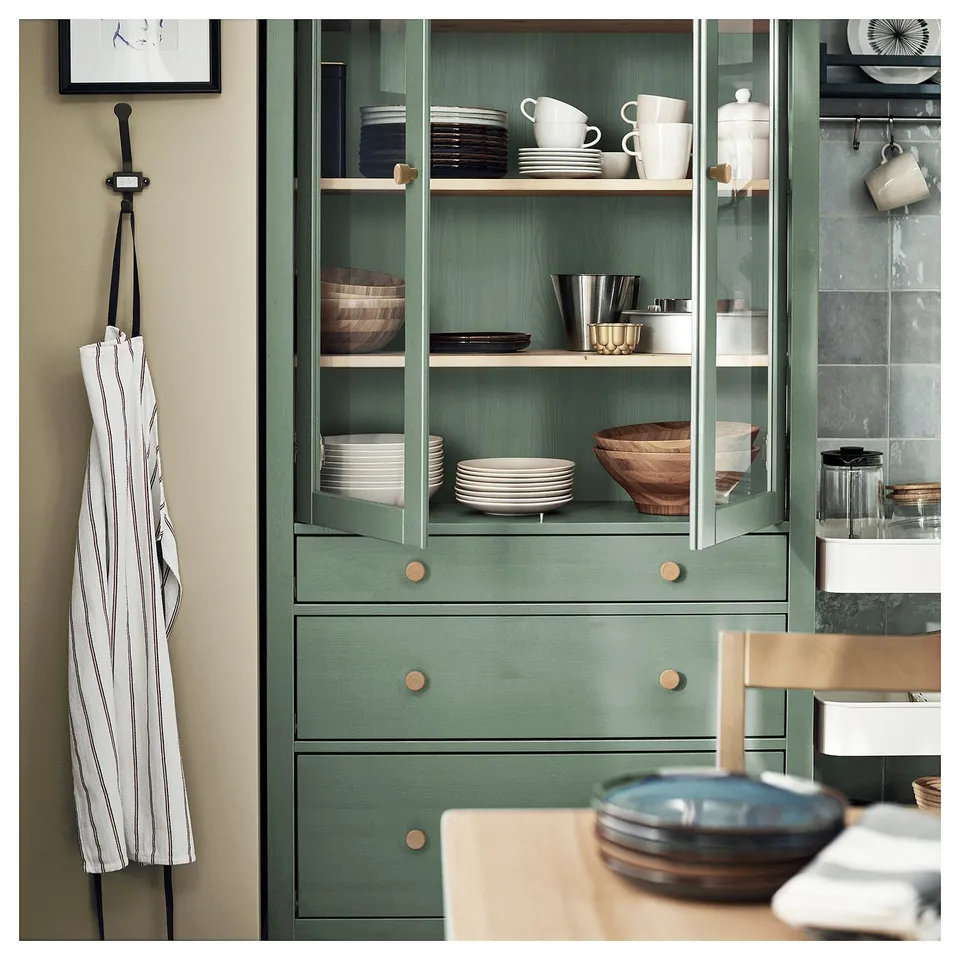 Green HEMNES cabinet with glass doors, wood shelves, white dishes, cups, and kitchen tools. Apron hangs nearby.