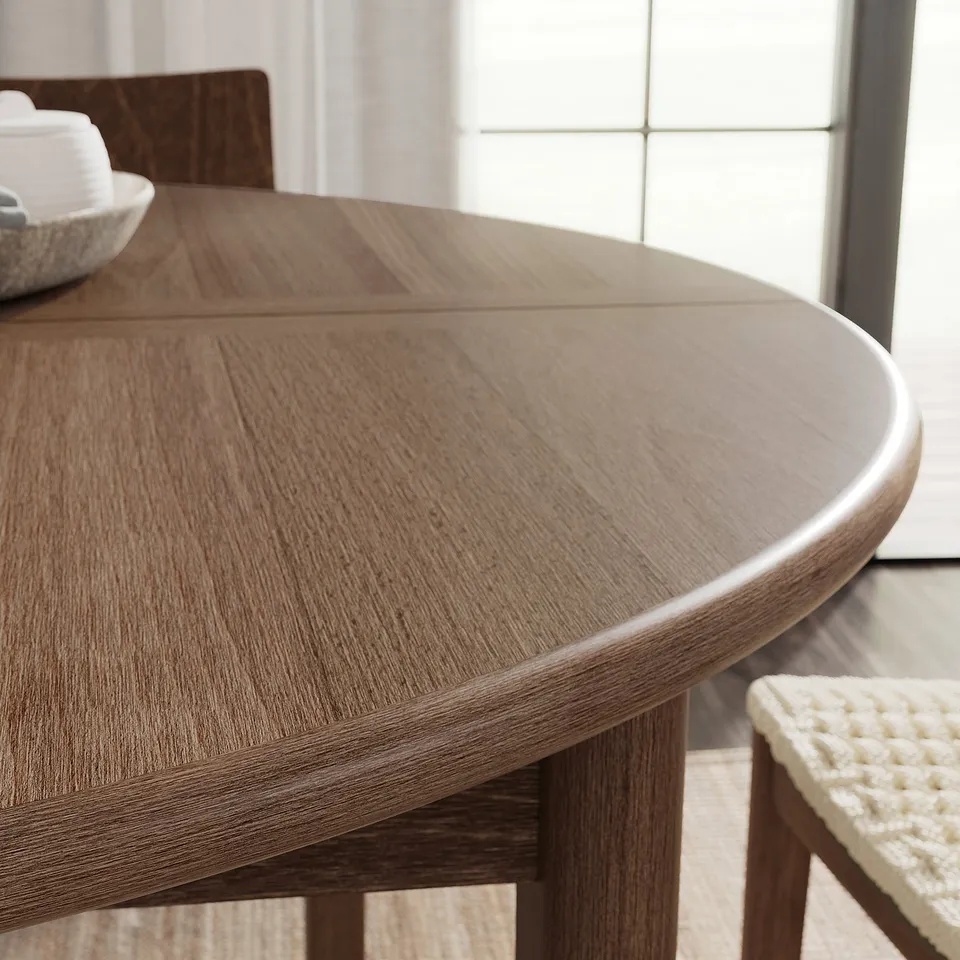 Modern round dining table with light brown finish, smooth edges, and a wooden texture. A chair with a woven seat is visible next to it.