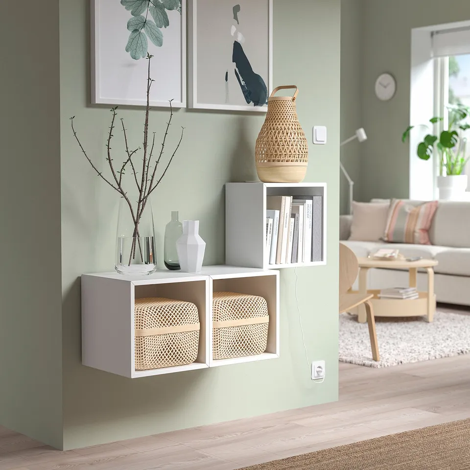 Minimalist living room with light green walls, white EKET shelves, baskets, and vases, natural light.