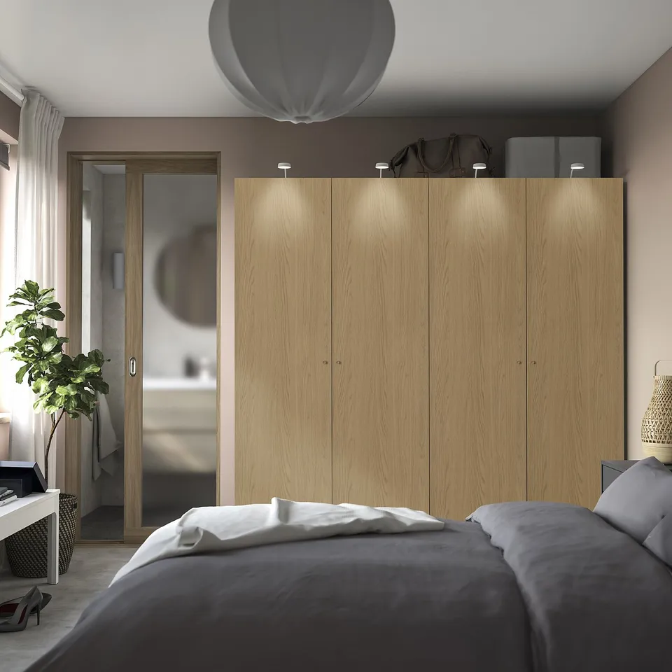 A modern bedroom with a large wooden PAX wardrobe and built-in lighting, positioned near a bed with grey bedding.