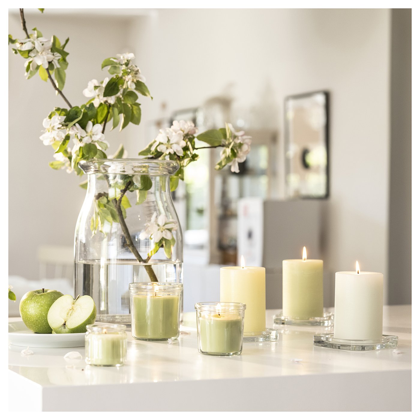 Cosy kitchen scene, apples, lit candles, and blooms. Apple-scented green candles beside apples in a jar.