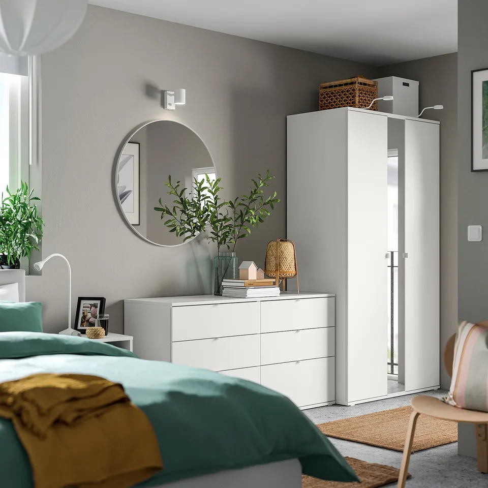 Modern bedroom with teal bedding, white furniture, round mirror, and green plant.
