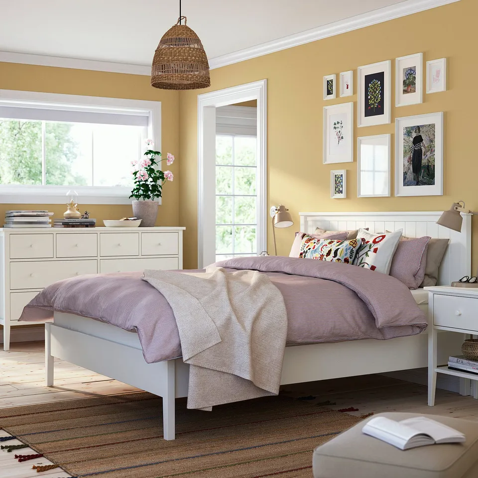 Yellow bedroom with white bed, pink blanket, chest, art, window, lamp, plant.
