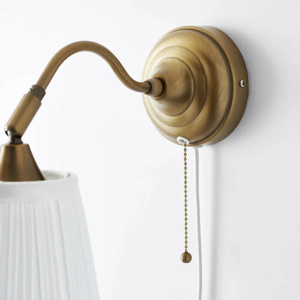 Gold wall-mounted lamp with adjustable arm and pull chain. White textured shade casts a soft light.