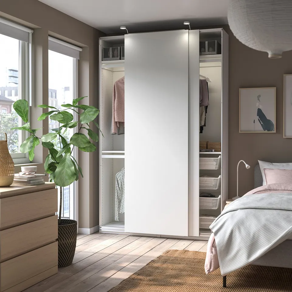 Modern bedroom a white PAX wardrobe with mirrored sliding doors, a bed with pink accents, a wooden dresser, and a large green plant by the window.