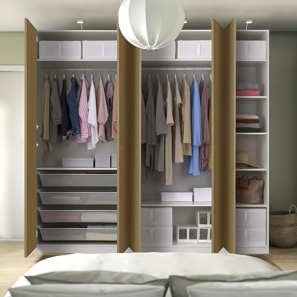 Open wardrobe displaying folded clothes, hangers, and storage boxes. A bed with white pillows in front, lit by a white pendant lamp.