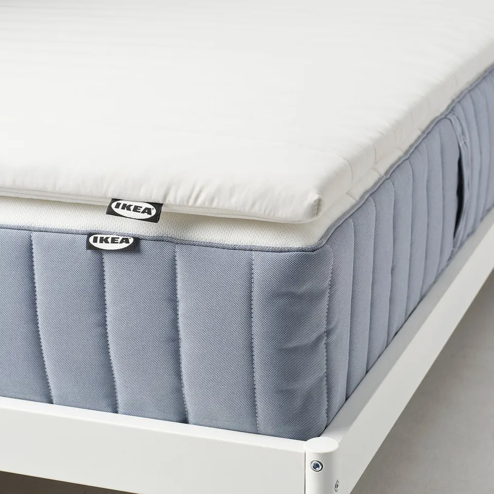 A thick, white mattress pad by IKEA, providing extra softness and cushioning for comfort. The pad a washable cover on a grey base.