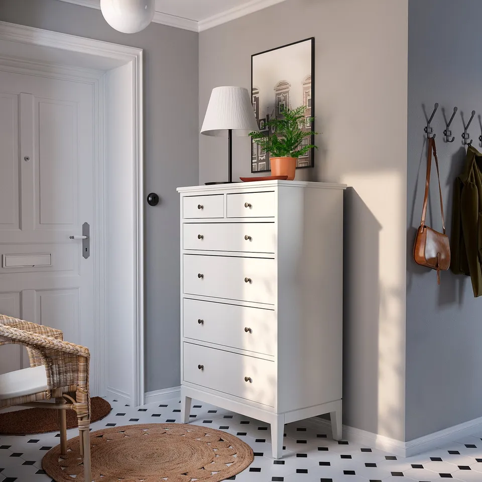 Modern entryway with white IDANÄS dresser, black hooks, coats, bag, lamp, plant, mirror, woven chair, geometric rug.