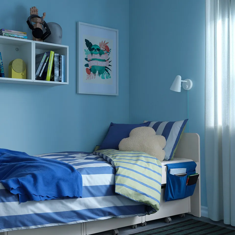 Kids bedroom with blue walls, white bed, striped bedding, cloud pillow, grey bed pocket with books, colourful shelf, picture, wall lamp.