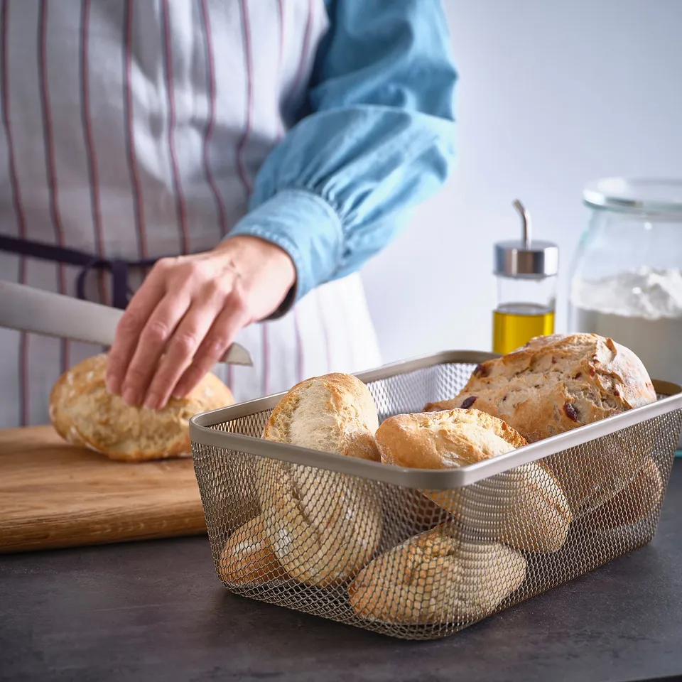 Person arranging bread in an IKEA 365+ mesh basket on a kitchen counter. The basket allows air circulation for better taste and less waste.