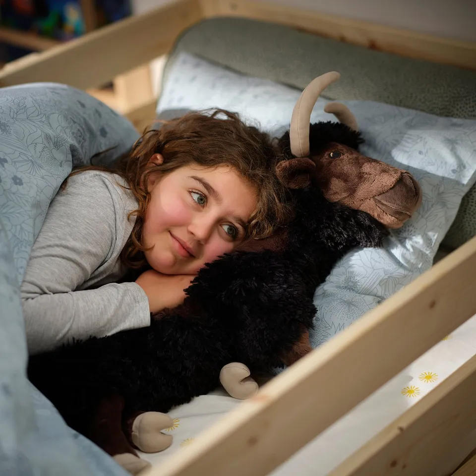 A girl snuggles a plush miner moose toy in a cosy bed, likely from the ALPTALL collection, evoking curiosity about himalayan wildlife.