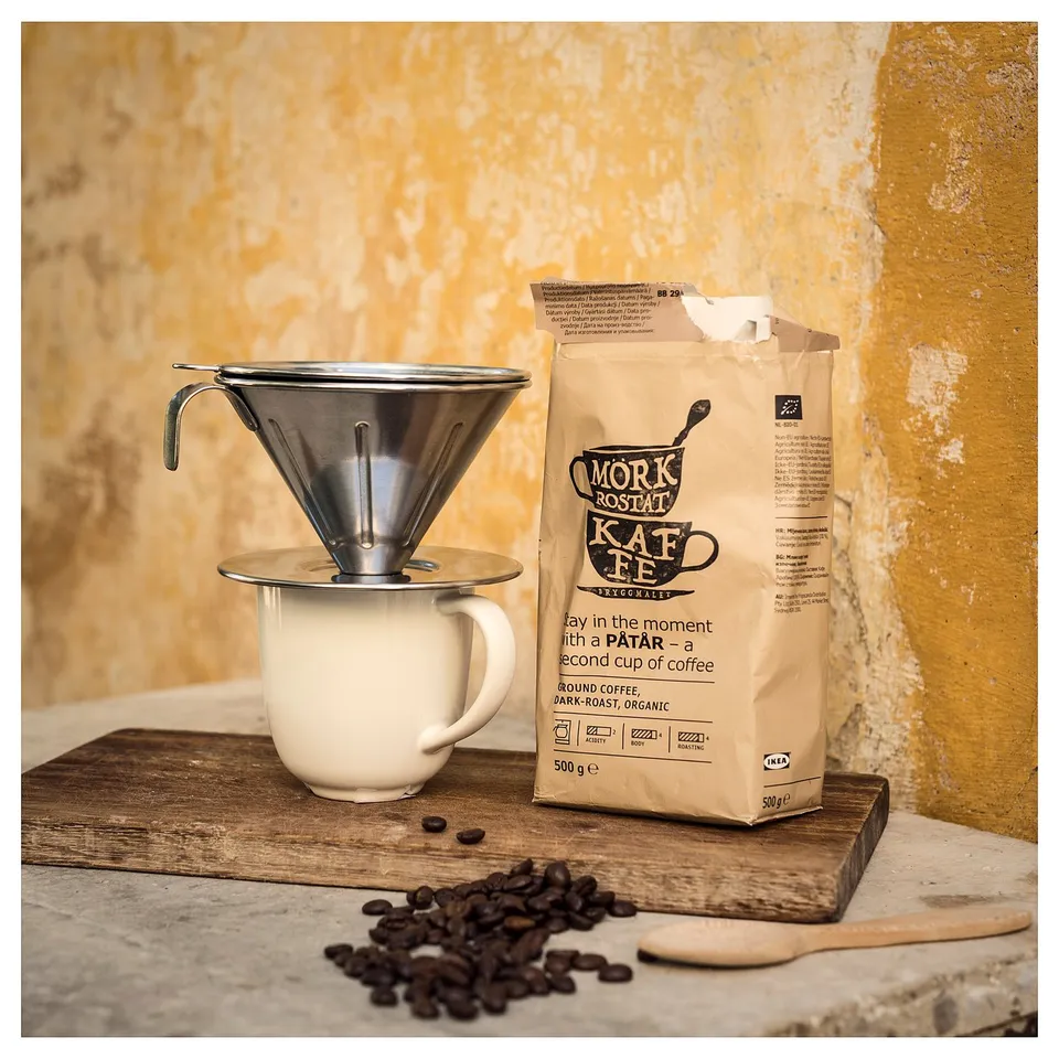 A coffee setup with a white enamel mug, metal pour-over philtre, and beige coffee bag on a wooden board.