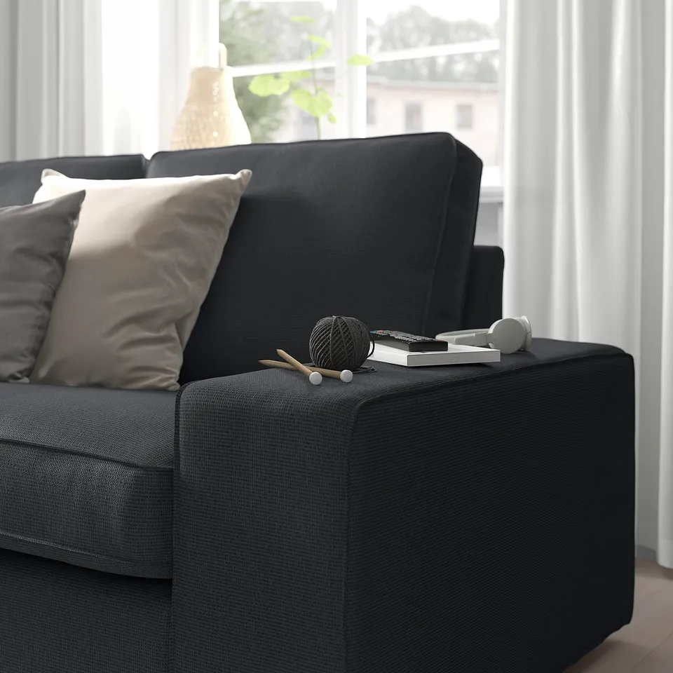 Modern living room with black KIVIK sofa, featuring pillows, remote, yarn and knitting needles on armrest, by window.