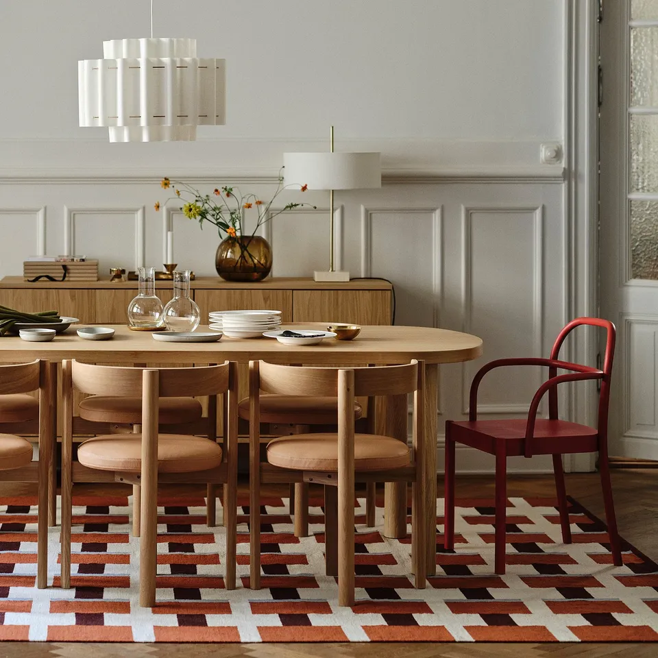 Modern dining area with round wooden table, six chairs, red accent chair, patterned rug, pendant light, sideboard with lamp, and vase with flowers.