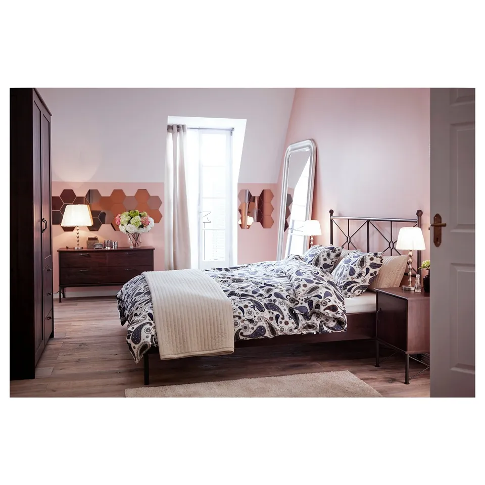 Elegant bedroom with metal bed, pink walls, honeycomb mirrors, wooden dresser, and bed lamps. Large window, cosy rug.