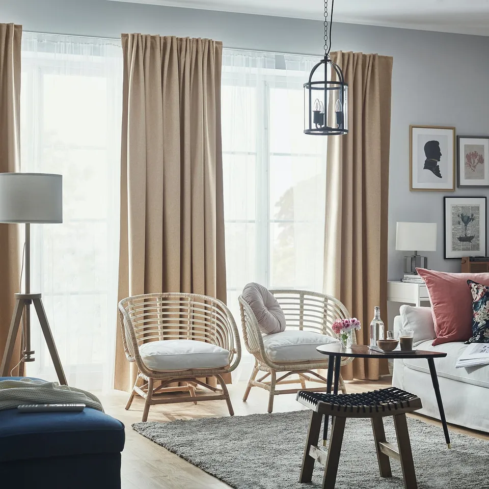 Room-darkening curtains frame wicker chairs, white sofa, and wooden coffee table. Grey rug and light wood floor complete cosy living room.