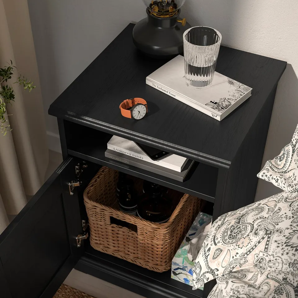 Dark bedside table with open drawer showing wicker basket and bottles. Tabletop holds books, glass, and watch.