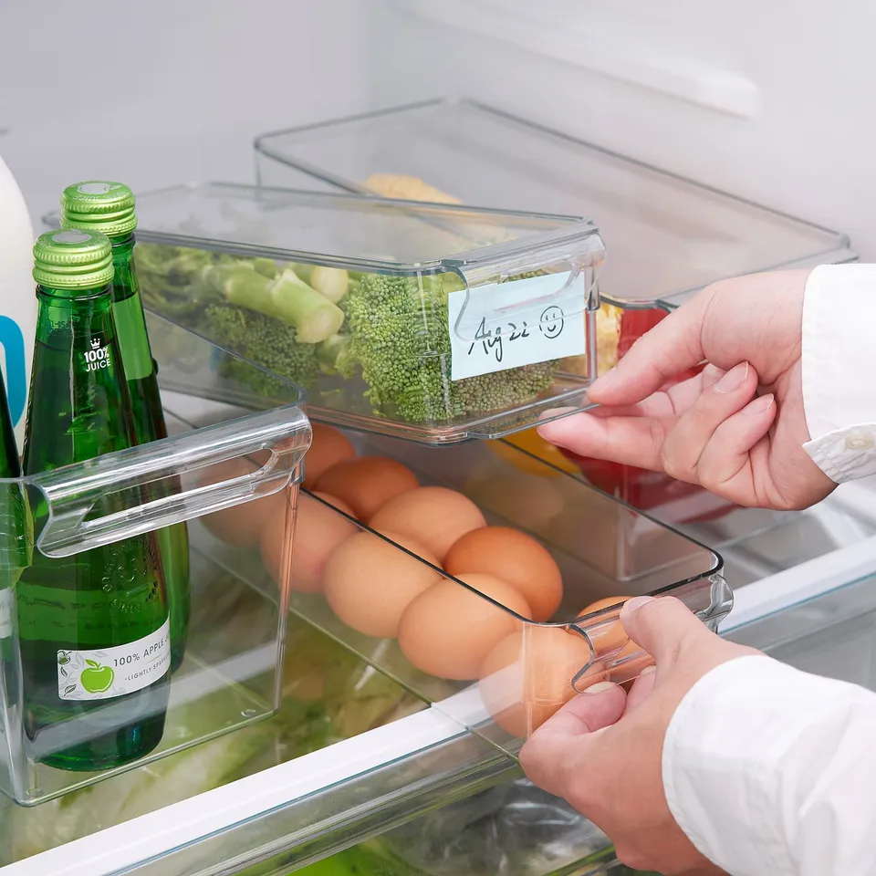Person organising fridge with transparent boxes. One box has broccoli, another has eggs. Boxes fit neatly on one shelf, maximising space.