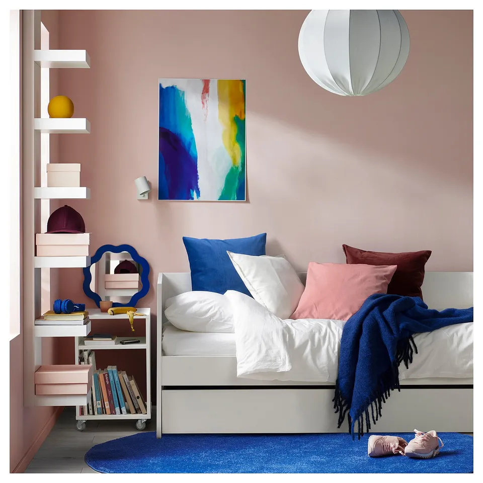 Cosy bedroom with a white day-bed, colourful pillows, and a blue rug. Artwork and hanging lamp on pink walls.