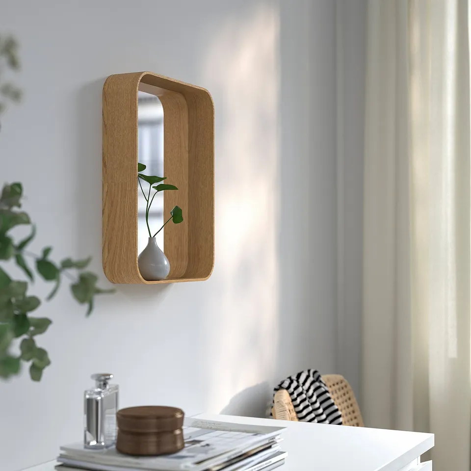 A wooden-framed mirror with shelf holds vase, mounted on a white wall.