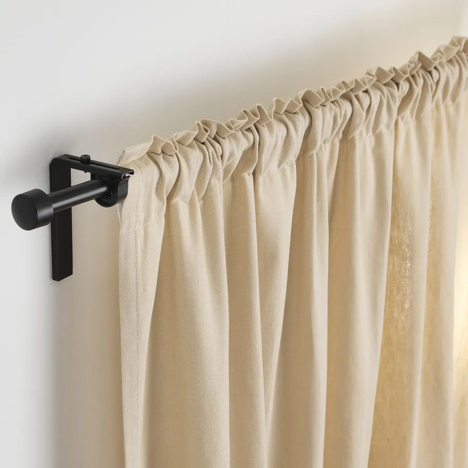 Black curtain rod with adjustable length and angled holder supporting beige curtains.