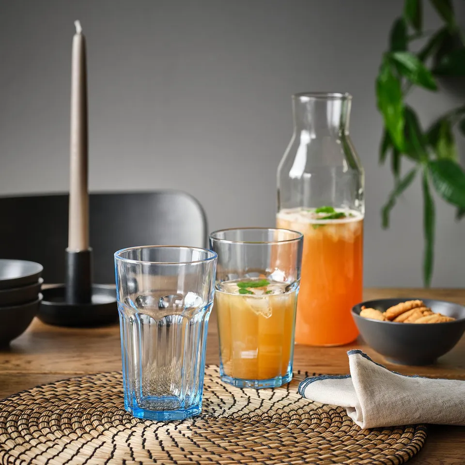 Clear glasses on woven mat, one filled with orange liquid, another empty, beside pitcher, candle, plant, snacks, and bowls.