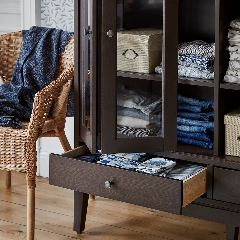 Organised cabinet with folded clothes, woven chair, & teal throws. Dark wood, glass doors.