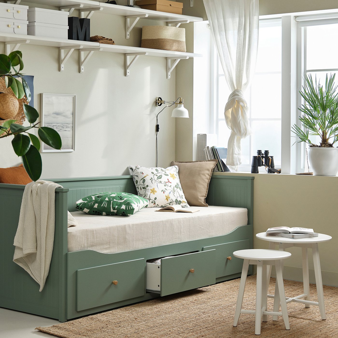 HEMNES grey day-bed sofa/bed with storage, surrounded by white shelves and furniture, with a window and plants.