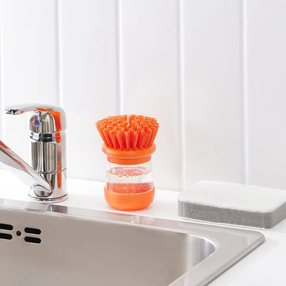 A stainless steel sink with an orange washing-up brush and soap dispenser next to a chrome tap. The brush holder dispenses soap onto dishes.