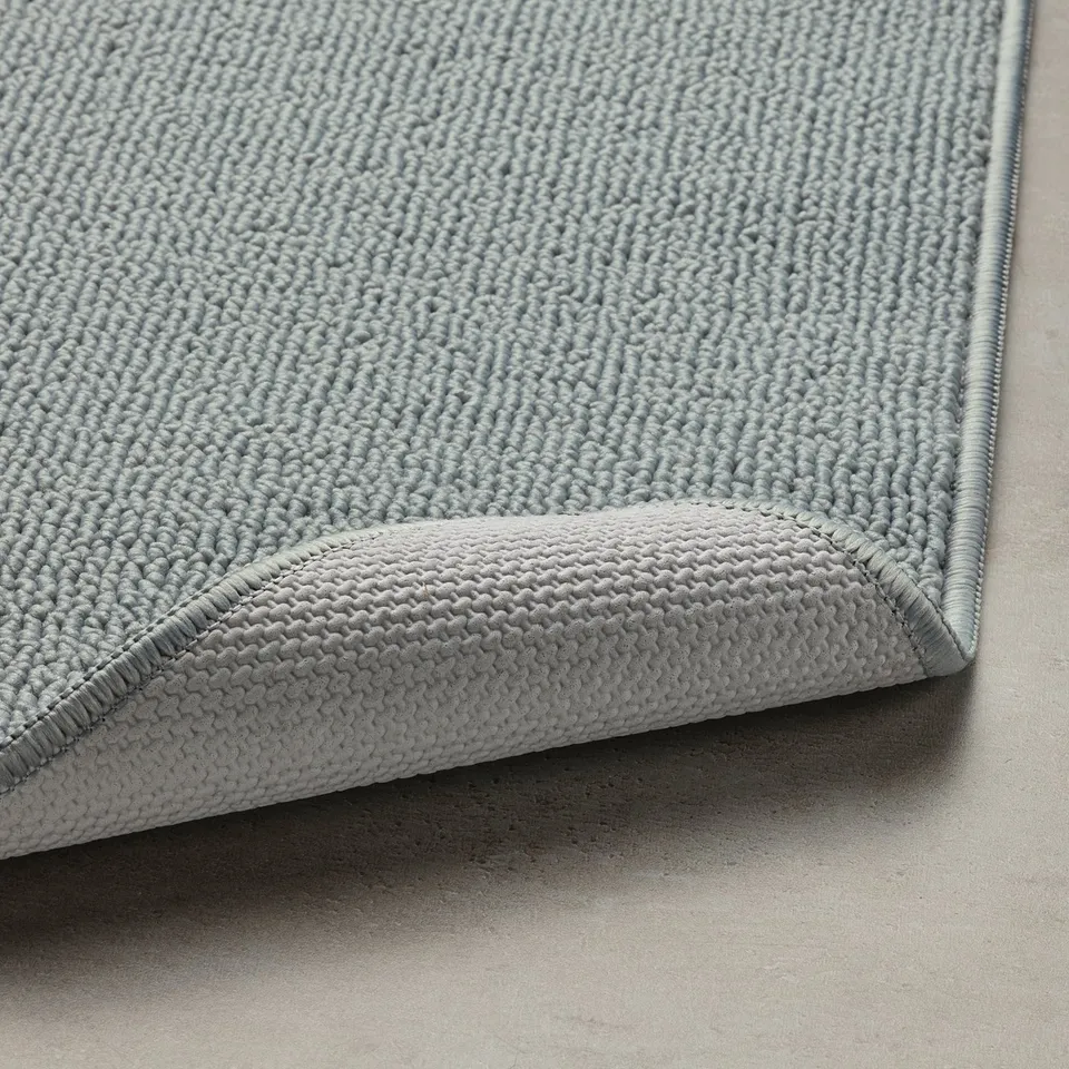 Light blue textured bath mat with a flatwoven surface, latex backing for stability, and rolled corner shown.