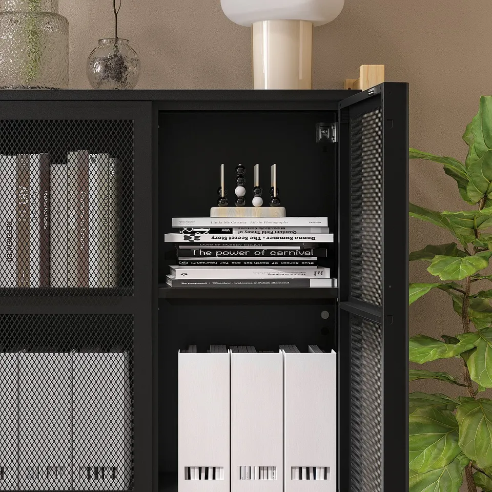 Black IVAR storage unit with open door revealing books and records. Mesh doors on either side, placed against wall with decor items.