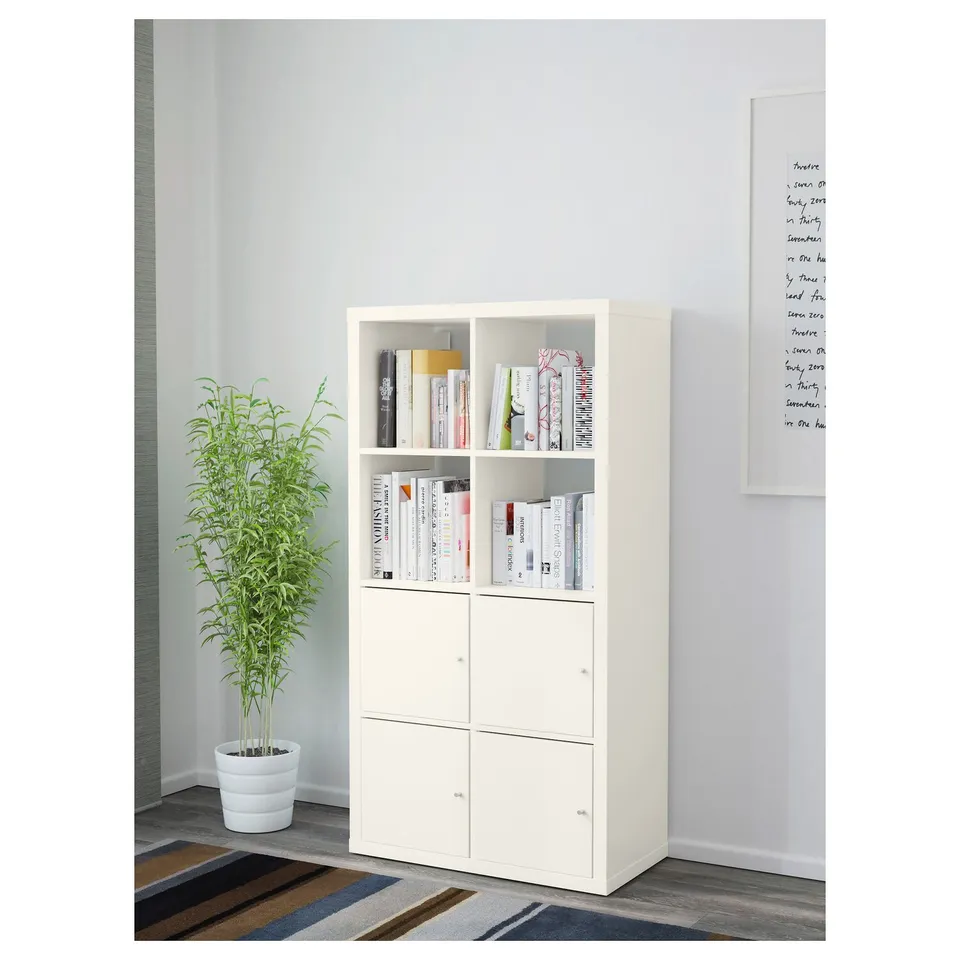 White KALLAX shelf with books, plant, and wall-mounted whiteboard.