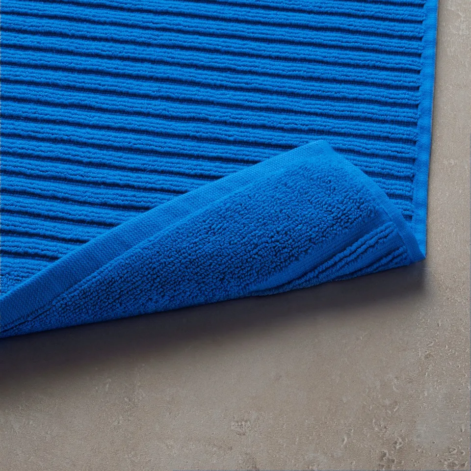 Bright blue bath mat on grey floor, folded edge showing thick, absorbent cotton.