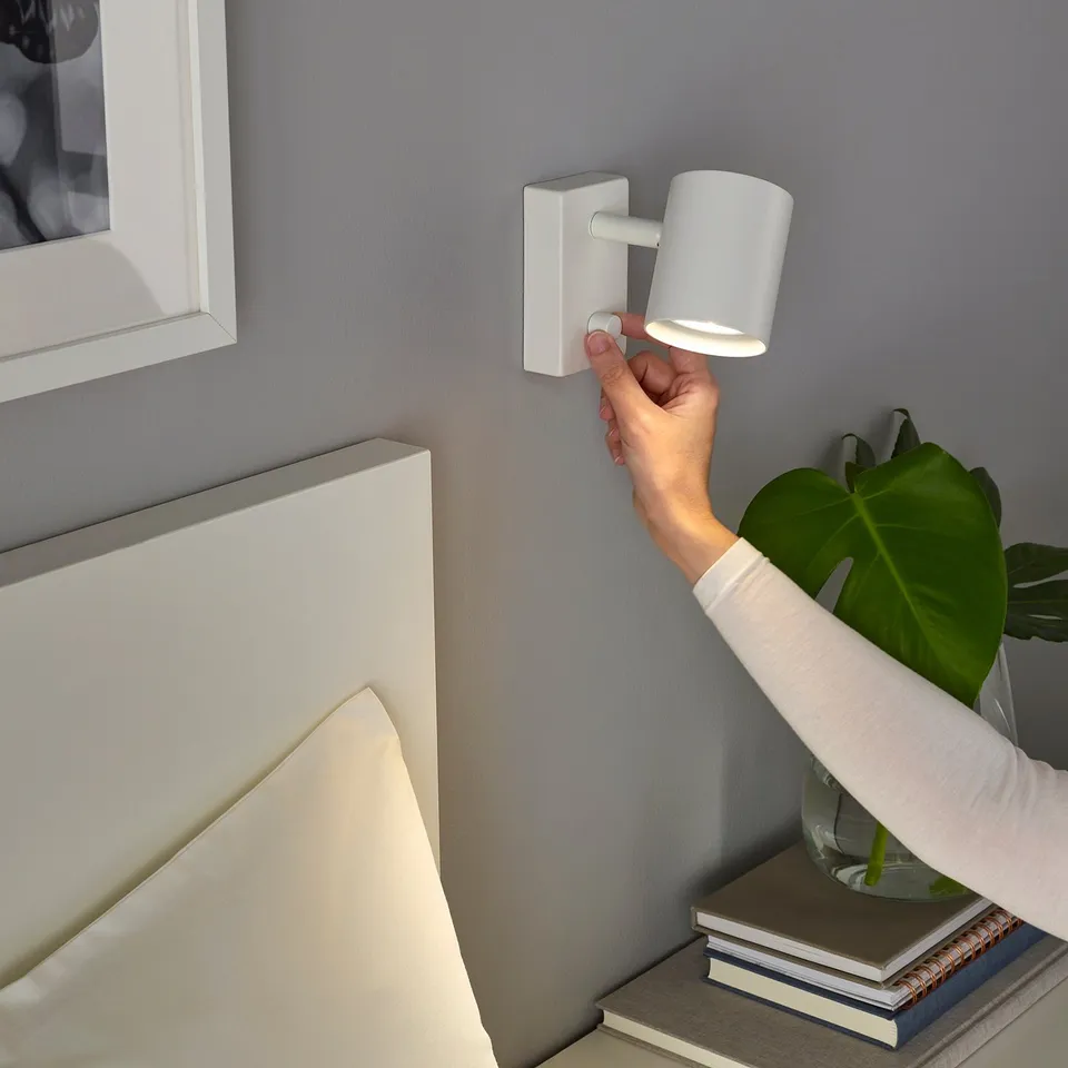Hand adjusts wall-mounted, dimmable white light fixture near bed.
