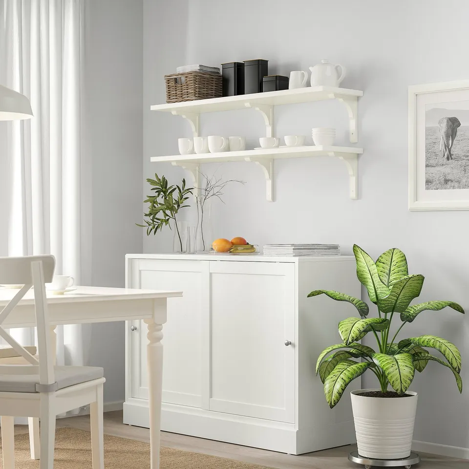 White kitchen corner with BERGSHULT shelf holding cups, pitchers, books, and a large plant beside a cabinet and table against a light grey wall.