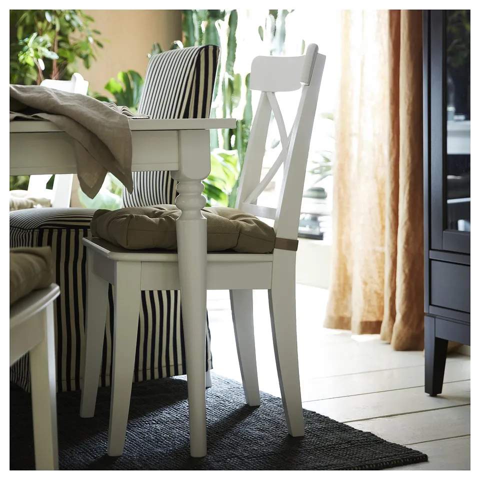 A white wooden chair with a striped cushion and a cosy rug beneath. The room is bright with a window and curtains in the background.