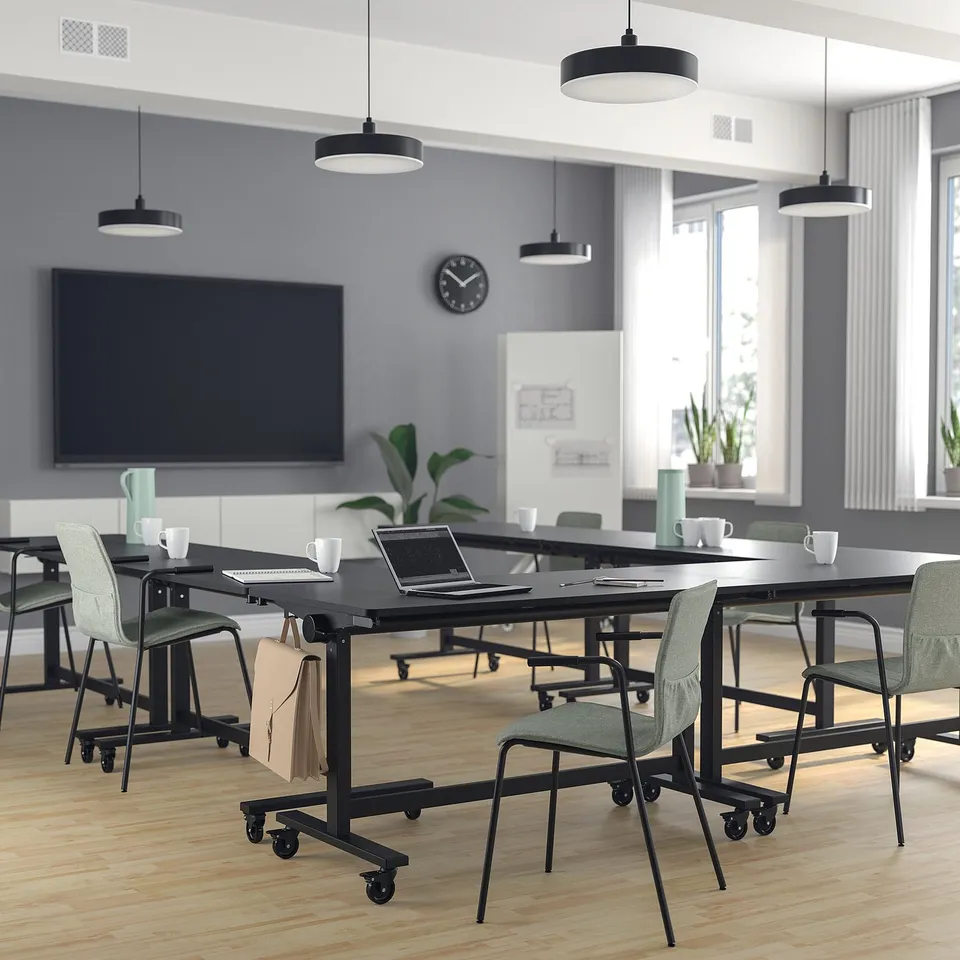 Modern conference room with MITTZON tables, foldable chairs. Cups, laptops, bags on tables. Large screen, clock. Big windows, lamps.