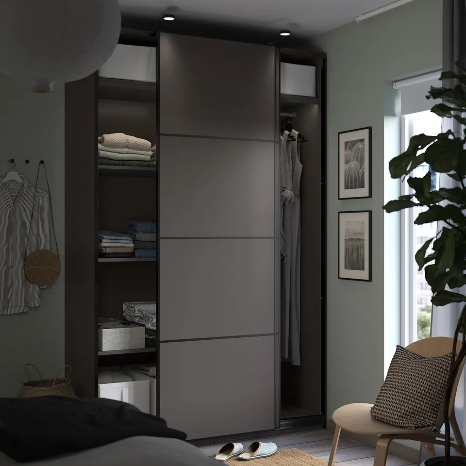 Modern bedroom PAX wardrobe in dark grey with sliding doors, shelves, and hanging rail. Wooden chair with cushion and plants decorate the space.