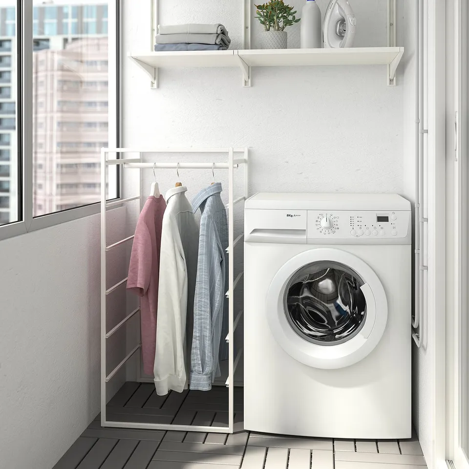 Small white JONAXEL clothes rack between washer & window, holding pink, white, & blue shirts, fitting neatly in compact room.