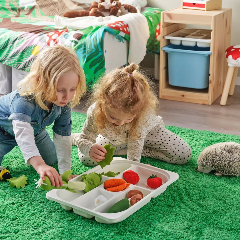 Two kids play with toy veggies in a TROFAST tray on a green rug.