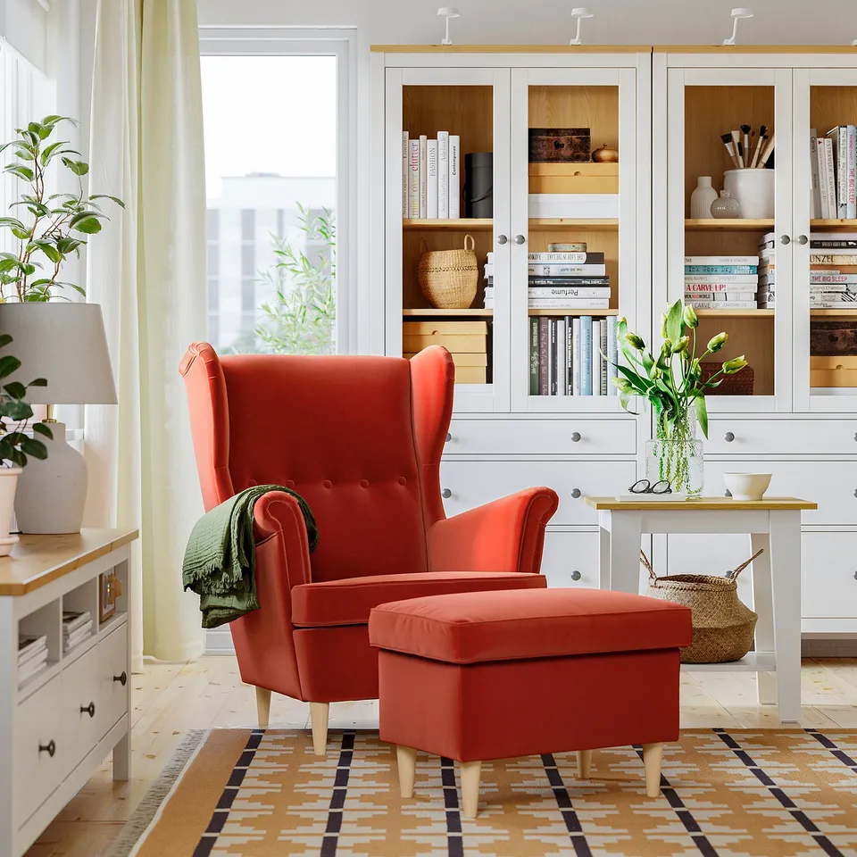 Bright red STRANDMON wing chair & ottoman on rug, beside white cabinet with books, near window.