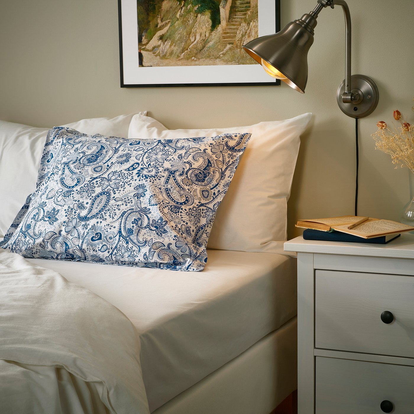 Bed scene with RODGERSIA blue paisley pillowcase, soft cotton-viscose blend, on a cream bed, beside a small bedside table with a lamp and books.