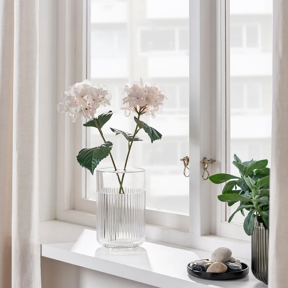 Windowsill decor: two vases, one with light pink flowers, another with green leaves. A bowl of smooth stones sits between them.