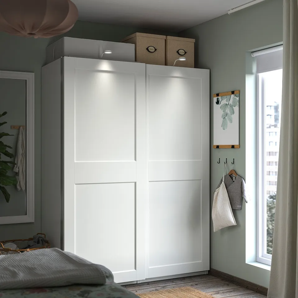 Modern white PAX wardrobe with sliding doors, top storage boxes, plant, clothes hangers near window.