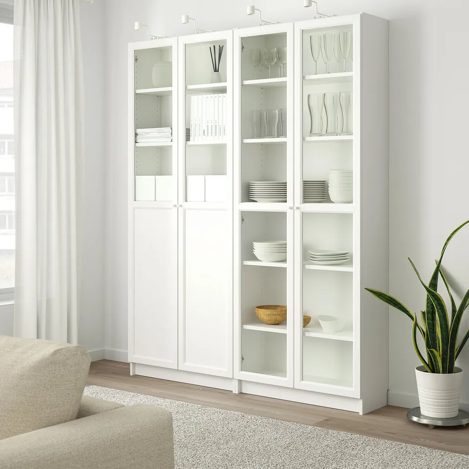 White BILLY bookcase with glass doors, storing dishes and decor, taller than a beige armchair in a bright room.
